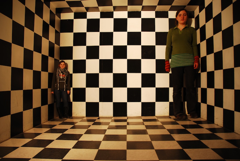 Two girls stand in a room with a striking black and white checkerboard pattern on the walls and floor, creating a surreal perspective.
